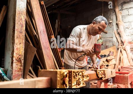 An Asian man focused on carpentry and painting in Bali, intricately working on a wooden piece. The workshop atmosphere showcases traditional skills an Stock Photo
