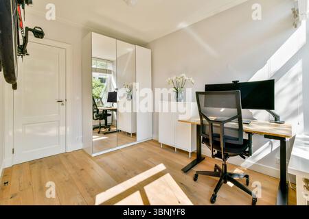 A stylish office space featuring a desk, computer setup, and a large mirror reflecting the room. Natural light enhances the ambiance, creating a produ Stock Photo