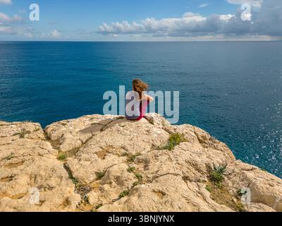 A young woman sits on rocky cliffs at Costa Blanca, Spain, gazing out at the expansive azure sea under a partly cloudy sky, capturing a serene moment Stock Photo