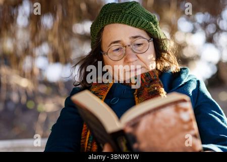 A middle-age woman dressed warmly in a coat, scarf, and hat, enjoys reading a book while sitting outdoors in a serene countryside setting during cold Stock Photo