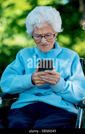 An elderly woman in a wheelchair is focused on using her smartphone outdoors. She appears engaged and content, wearing a cozy blue jacket against a bl Stock Photo