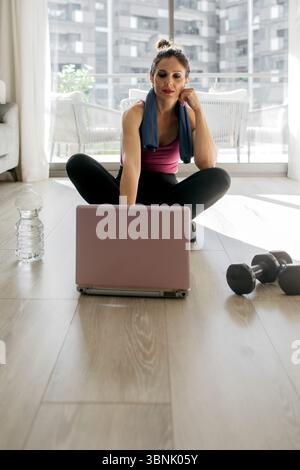 Woman working out at home, sitting on the floor, using a laptop for a guided fitness session. Nearby are a water bottle and dumbbells, indicating an e Stock Photo