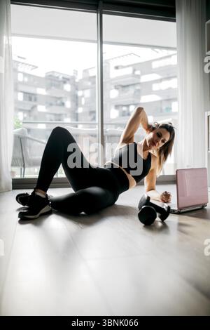 Woman in athletic wear training at home, using a laptop for guidance. She is working out on a wooden floor, with dumbbells nearby, emphasizing fitness Stock Photo