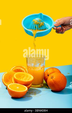 Cropped unrecognizable hand holding a vibrant blue juicer being used to squeeze fresh orange juice into a glass set against a contrasting yellow and b Stock Photo