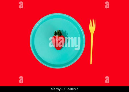 A vivid image featuring a single ripe strawberry placed on a blue plate with a yellow plastic fork beside it, all set against a striking red backgroun Stock Photo