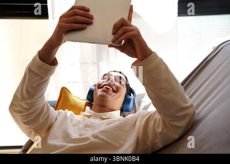 A young Arab man relaxes on a couch, wearing blue headphones and holding a tablet. He smiles as he enjoys his leisure time in a bright, comfortable se Stock Photo