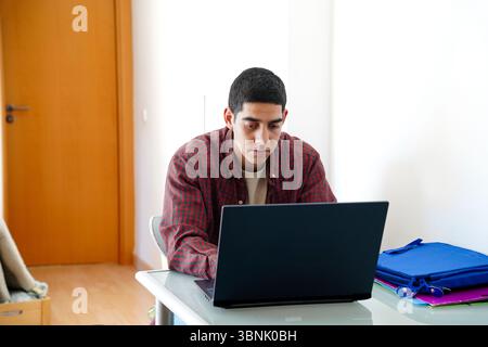 Young Arab man in casual attire working on a laptop at home. He is engaged in his daily routine, with a thoughtful expression, concentrating on the sc Stock Photo