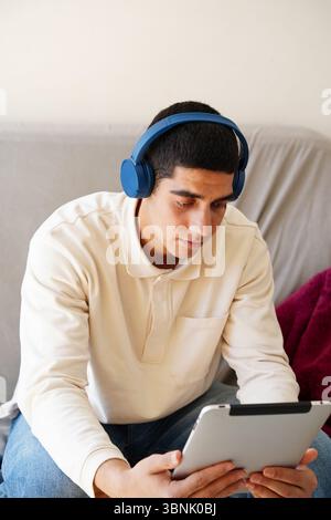 A young Arab man sits on a couch, focused on his tablet. Wearing blue headphones, he seems engaged with digital content, reflecting a modern lifestyle Stock Photo