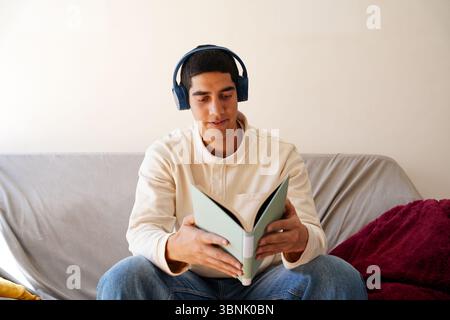 A young Arab man listens to music with headphones while reading a book on the couch, embracing a moment of relaxation in his daily routine at home Stock Photo