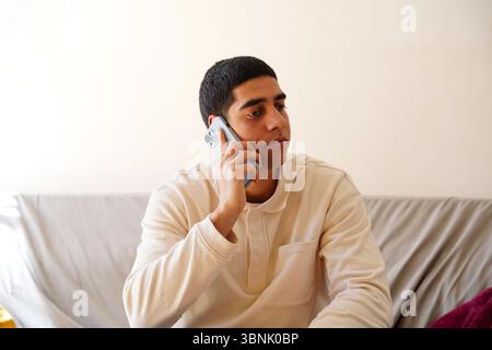 Young Arab man in a casual setting, holding a smartphone and engaging in conversation. He seems thoughtful and focused, sitting comfortably on a sofa Stock Photo