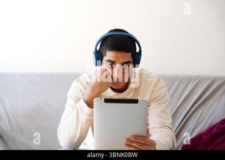 Young Arab man wearing headphones, absorbed in his daily routine, as he attentively engages with content on a tablet while seated on a comfortable sof Stock Photo