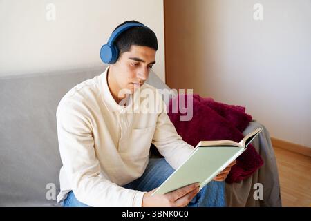 A young Arab man sits on a couch at home, wearing headphones and reading a book. He is focused and relaxed, enjoying a moment of leisure in his daily Stock Photo