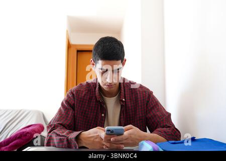 A young Arab man is focused on his smartphone during his daily routine, wearing a casual red plaid shirt. The indoor setting adds a sense of everyday Stock Photo