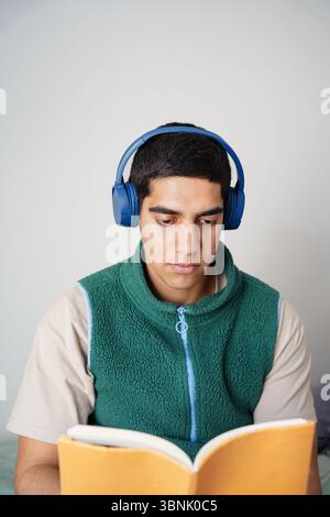 Young Arab man sits casually with headphones on, engaged in reading a book, showcasing a moment of learning and relaxation in his daily routine. Moder Stock Photo