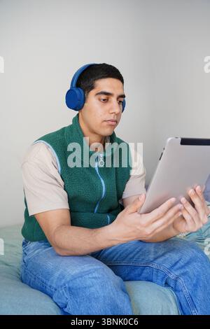A young Arab man is sitting on a bed, deeply engaged with his tablet while wearing blue headphones. He is dressed casually, absorbed in his daily rout Stock Photo