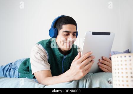 Young Arab man lying on a couch with headphones, engaged in digital content on a tablet. Capturing modern lifestyle and daily routine, focused on rela Stock Photo