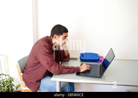 Young Arab man wearing a red checkered shirt, sitting at a desk with a laptop. He looks focused and thoughtful, surrounded by supplies, in a bright ro Stock Photo