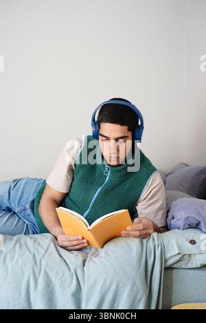 A young Arab man relaxes on his bed, wearing headphones and reading a book. Dressed casually in a green sleeveless jacket and jeans, he enjoys a peace Stock Photo