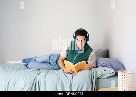 A young Arab man lies comfortably on his bed, absorbed in a book, while wearing headphones that offer a serene escape during his daily routine at home Stock Photo