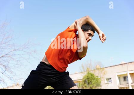 Young Arab man in casual sportswear stretching outdoors on a sunny day. He is engaging in a fitness routine, demonstrating flexibility and focus again Stock Photo