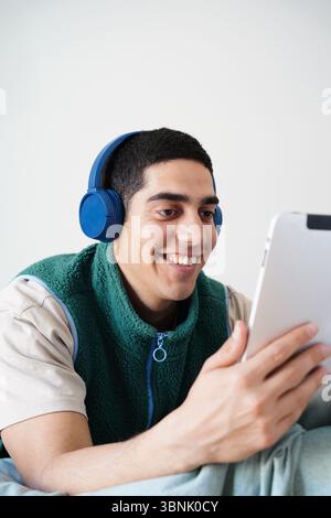 Young Arab man enjoying his daily routine, wearing headphones while engaging with a tablet. He is relaxed and smiling, immersed in a digital experienc Stock Photo