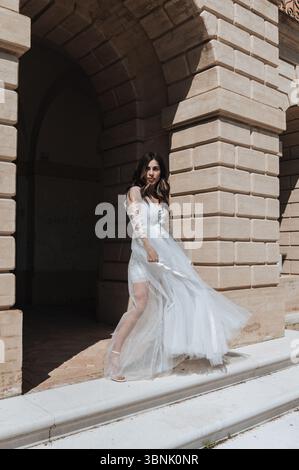 A bride in a flowing white dress stands gracefully by a stone archway, capturing the elegance and charm of a wedding day. Sunlight highlights her intr Stock Photo