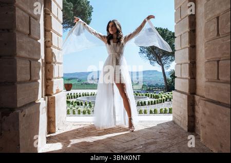A bride in a flowing white dress poses elegantly in a picturesque outdoor setting. The serene landscape, with lush greenery and blue skies, creates an Stock Photo