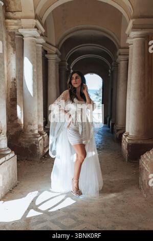 A bride in a stunning white dress poses gracefully in a rustic corridor, with light filtering through the archways. The picturesque setting enhances t Stock Photo