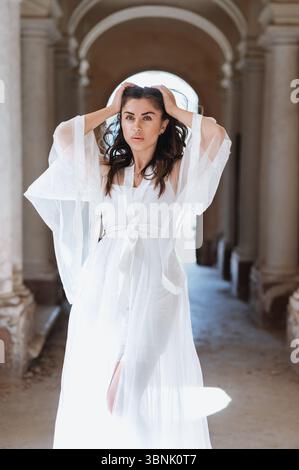 A bride in a stunning white gown poses gracefully in a historic corridor, exuding elegance and confidence on her special day. The sunlight highlights Stock Photo