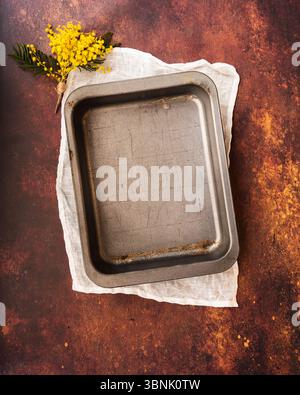 An empty rectangular baking pan sits atop a white cloth on a textured, warm-toned surface. A small bouquet of yellow flowers adds a touch of color and Stock Photo
