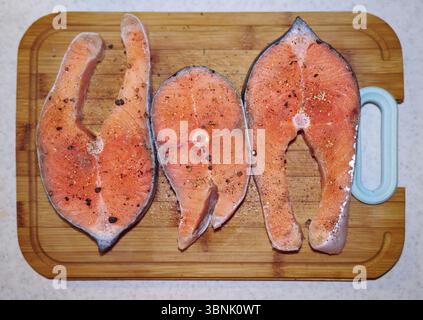 Three raw salmon steaks are neatly arranged on a wooden cutting board, showcasing a vibrant orange hue with specks of seasoning. Perfect for culinary Stock Photo