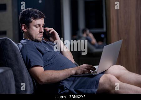 A man comfortably sits on a couch, engaged in conversation on a cell phone while simultaneously using his laptop for various tasks Stock Photo