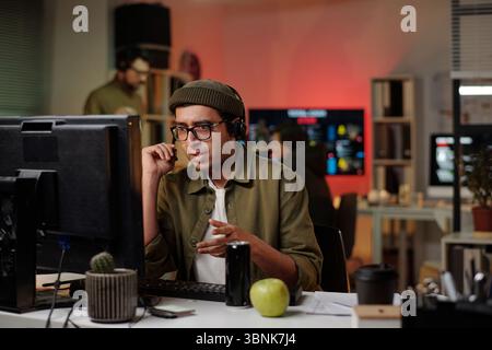 Young man wearing headset sitting at desk operating computer while speaking into microphone in scam call center with coworkers in background Stock Photo