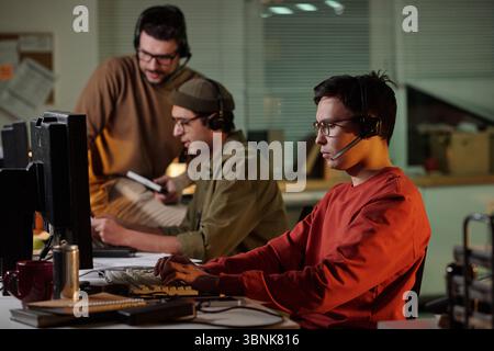 Three young adult men wearing headsets operating computers in office setting, collaborating during scam call center operation Stock Photo