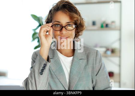 Portrait of a confident pretty brazilian businesswoman in blazer adjusting her glasses and smiling. Young successful lady posing at office in formal attire with self-assured expression Stock Photo