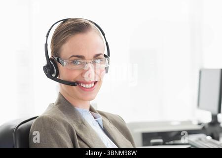 Customer service agent wearing headset and glasses, speaking at office desk by window with monitor Stock Photo