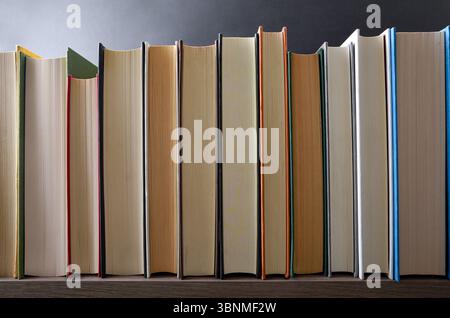 Row of books showing the front edge on a wooden shelf and dark isolated background. Front view. Stock Photo