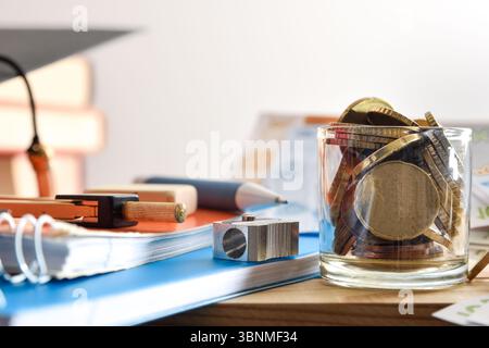 Detail and concept of the cost of school supplies with a jar full of coins  and school supplies and books around it on a table and isolated white back Stock Photo