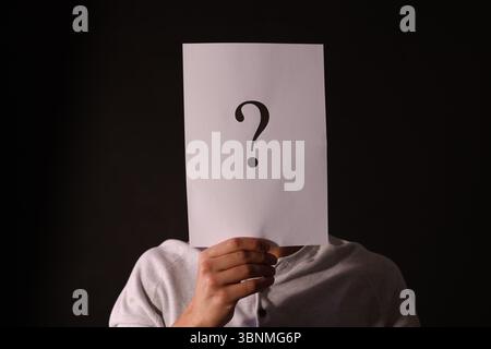 Man hiding behind paper sheet with question mark on dark background Stock Photo