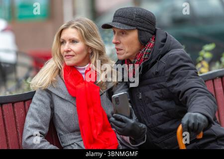 An elderly man and a woman are sitting on a bench, talking and looking away. They are in warm clothes. Stock Photo