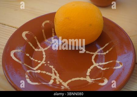 A round orange fruit sits on a brown ceramic plate, which features an abstract white handprint design. It rests on a light wooden surface. Stock Photo