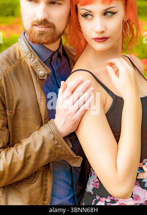 A man in a brown leather jacket embraces a woman with red hair, who is wearing a black top and floral skirt. Stock Photo