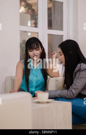 Two young women are engaged in light conversation over coffee, in a comfortable and relaxing interior setting. The duo shares a pleasant moment of soc Stock Photo