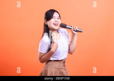 A happy asian woman is singing into a microphone with an expressive and joyful look, isolated on orange background Stock Photo