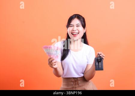 A happy young woman smiles while holding money and a car key, symbolizing financial success and new car ownership, isolated on orange background. Stock Photo