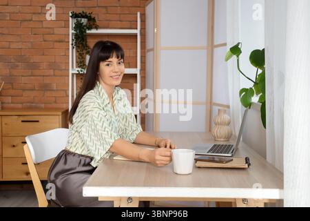 Beautiful woman with cup of coffee and laptop on table at home Stock Photo
