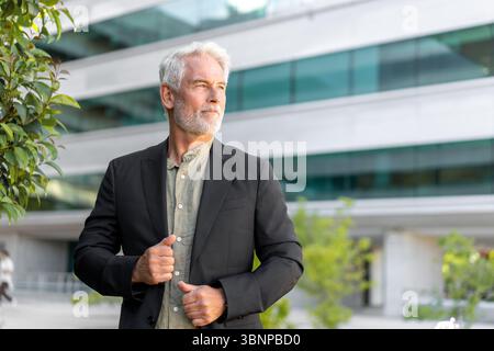 Senior businessman in formal suit standing outside modern office building adjusting blazer. Confident and focused in natural daylight setting Stock Photo