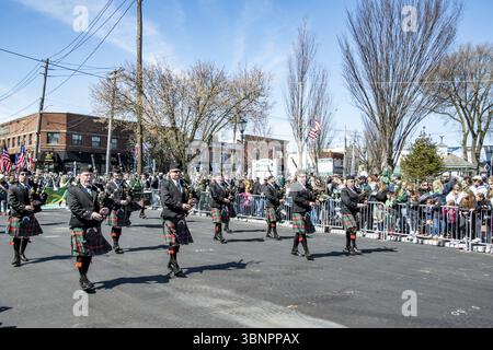 Lindenhurst, New York, USA, March 30, 2024 - Group Of Bagpipers Wearing Traditional Scottish Kilts And Black Jackets March Down A Sunlit Street During Stock Photo