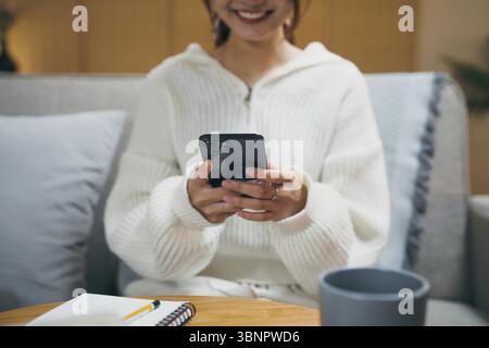 Happy asian woman using mobile phone and receiving good news on sofa at living room Stock Photo
