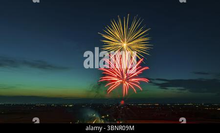 Bright Fireworks Exploding In The Night Sky With Vibrant Red And Yellow Colors Over A Cityscape With Distant Lights, Set Against A Dark Blue And Green Stock Photo